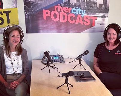 Two women are sitting at a table with microphones in front of a river city podcast sign.