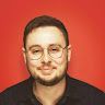 A man with glasses and a beard is smiling in front of a red background.
